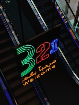 A neon sign featuring large, brightly lit numbers '3, 2, 1' in green, red, and blue respectively. The word 'Welcome' is displayed in yellow neon below the numbers, along with Arabic script in the same color. The sign is positioned against the backdrop of escalators, creating a dynamic visual contrast.