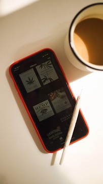 A smartphone with a red case displaying a reading library with books related to cannabis. A rolled joint is placed on top of the phone, and next to it is a cup of coffee.