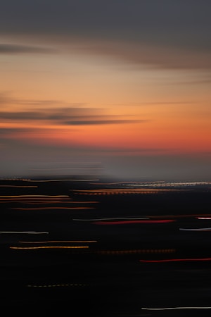 A blurred cityscape at dusk with a gradient sky transitioning from orange to dark blue. The lights from the city create streaks of light, suggesting movement and vibrancy.