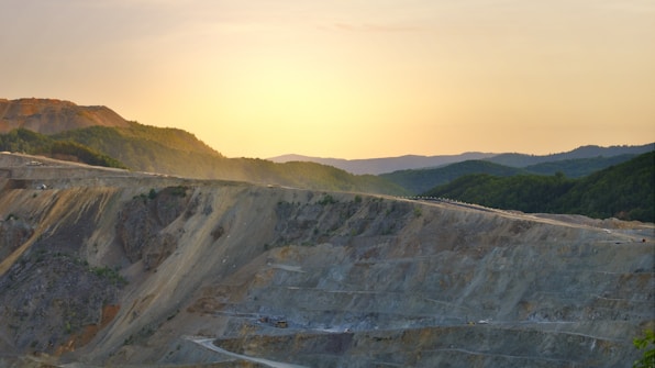 A vast open-pit mine extends across the foreground, with terraced layers cutting through rocky terrain. In the background, rolling hills covered in dense green forest gradually rise, while the sun sets on the horizon, casting a warm golden glow over the landscape.
