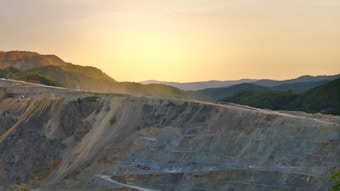 A vast open-pit mine extends across the foreground, with terraced layers cutting through rocky terrain. In the background, rolling hills covered in dense green forest gradually rise, while the sun sets on the horizon, casting a warm golden glow over the landscape.