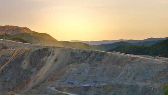 A vast open-pit mine extends across the foreground, with terraced layers cutting through rocky terrain. In the background, rolling hills covered in dense green forest gradually rise, while the sun sets on the horizon, casting a warm golden glow over the landscape.