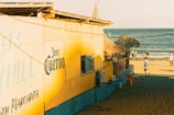 A vibrant beach hut with a yellow facade and thatched roof sits next to the shoreline. The building has a prominent 'Jose Cuervo' sign and displays hanging clothes outside. Several people walk along the sandy beach near the ocean, with a clear blue sky overhead.