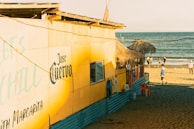 A vibrant beach hut with a yellow facade and thatched roof sits next to the shoreline. The building has a prominent 'Jose Cuervo' sign and displays hanging clothes outside. Several people walk along the sandy beach near the ocean, with a clear blue sky overhead.