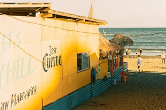 A vibrant beach hut with a yellow facade and thatched roof sits next to the shoreline. The building has a prominent 'Jose Cuervo' sign and displays hanging clothes outside. Several people walk along the sandy beach near the ocean, with a clear blue sky overhead.