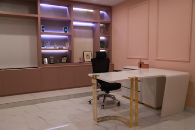 A modern office setup featuring a white L-shaped desk with gold accents and a black ergonomic chair. The wall behind the desk has built-in shelves illuminated with purple lighting, showcasing various trophies, awards, and decorative items. The decor includes blush pink walls and white marble flooring with gray veins. The overall design is minimalist and contemporary.