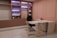 A modern office setup featuring a white L-shaped desk with gold accents and a black ergonomic chair. The wall behind the desk has built-in shelves illuminated with purple lighting, showcasing various trophies, awards, and decorative items. The decor includes blush pink walls and white marble flooring with gray veins. The overall design is minimalist and contemporary.