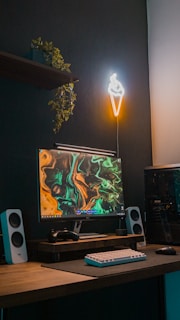 A modern computer setup features a sleek desktop display with an abstract, colorful wallpaper in greens and oranges. A white mechanical keyboard and black gaming mouse rest on a wooden desk. Above, a neon light shaped like an ice cream cone illuminates the dark, textured wall. A leafy plant hangs from the shelf above, adding a touch of nature.