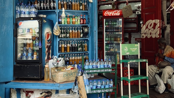 A small shop is filled with various bottled drinks, including Pepsi and Coca-Cola, displayed in coolers and on shelves. Water bottles are stacked on counters and in crates. A man sits nearby, wearing a mix of colorful and subdued clothing, adding a lived-in feel to the scene.