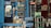 A small shop is filled with various bottled drinks, including Pepsi and Coca-Cola, displayed in coolers and on shelves. Water bottles are stacked on counters and in crates. A man sits nearby, wearing a mix of colorful and subdued clothing, adding a lived-in feel to the scene.