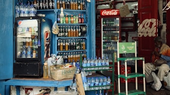 A small shop is filled with various bottled drinks, including Pepsi and Coca-Cola, displayed in coolers and on shelves. Water bottles are stacked on counters and in crates. A man sits nearby, wearing a mix of colorful and subdued clothing, adding a lived-in feel to the scene.