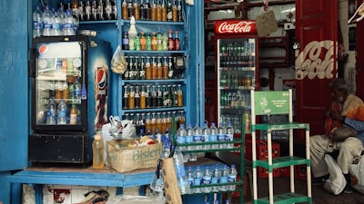 A small shop is filled with various bottled drinks, including Pepsi and Coca-Cola, displayed in coolers and on shelves. Water bottles are stacked on counters and in crates. A man sits nearby, wearing a mix of colorful and subdued clothing, adding a lived-in feel to the scene.
