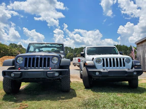 Two Jeep vehicles are parked on a grassy area under a bright blue sky with scattered clouds. The vehicle on the left is gray, while the one on the right is white. There is an American flag visible to the right of the vehicles, and some trees and a building are in the background.