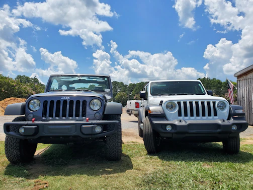 Two Jeep vehicles are parked on a grassy area under a bright blue sky with scattered clouds. The vehicle on the left is gray, while the one on the right is white. There is an American flag visible to the right of the vehicles, and some trees and a building are in the background.