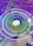 A colorful and futuristic digital artwork featuring a central logo surrounded by concentric neon rings. The background consists of a gradient of squares in purple and blue tones, with floating geometric shapes and a scattering of small lights.