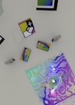 An array of abstract objects and colorful geometric patterns on a flat surface. The artwork includes a large square filled with pixelated, iridescent colors and surrounding shapes resembling books or frames with abstract designs.