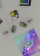 An array of abstract objects and colorful geometric patterns on a flat surface. The artwork includes a large square filled with pixelated, iridescent colors and surrounding shapes resembling books or frames with abstract designs.