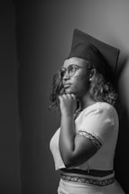 A black and white photograph of a person wearing a graduation cap and glasses, looking thoughtfully to the side. The subject has shoulder-length hair and is dressed in a traditional outfit with patterned trim. The background is plain, emphasizing the contemplative expression on the person's face.