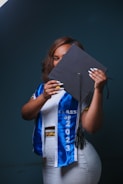 A person holding a graduation cap partially covering their face, wearing a white outfit with a blue graduation sash that reads 'Class of 2023'. The background is dark, creating a contrast with the brightly colored clothes.