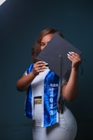 A person holding a graduation cap partially covering their face, wearing a white outfit with a blue graduation sash that reads 'Class of 2023'. The background is dark, creating a contrast with the brightly colored clothes.