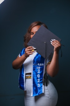 A person holding a graduation cap partially covering their face, wearing a white outfit with a blue graduation sash that reads 'Class of 2023'. The background is dark, creating a contrast with the brightly colored clothes.