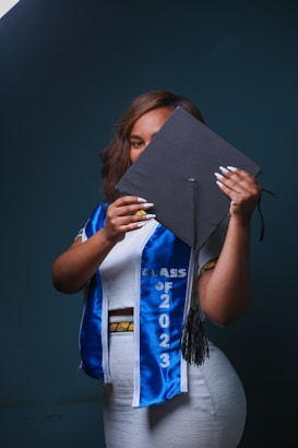 A person holding a graduation cap partially covering their face, wearing a white outfit with a blue graduation sash that reads 'Class of 2023'. The background is dark, creating a contrast with the brightly colored clothes.