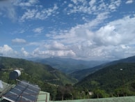 A vast mountainous landscape with lush green hills under a sky filled with scattered clouds. Solar panels and a satellite dish are installed on a structure in the foreground, overlooking the expansive view.