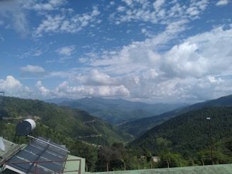 A vast mountainous landscape with lush green hills under a sky filled with scattered clouds. Solar panels and a satellite dish are installed on a structure in the foreground, overlooking the expansive view.