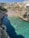 A picturesque beach scene with clear turquoise water surrounded by cliffs and a crowded sandy beach filled with people sunbathing and swimming. Colorful umbrellas dot the beach, and the backdrop features historic stone buildings.