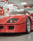 A vibrant red sports car is prominently displayed in what appears to be a showroom with reflective surfaces and bright overhead lighting. Two figures dressed in red racing suits and helmets stand in the background, adding to the high-performance and racing theme.