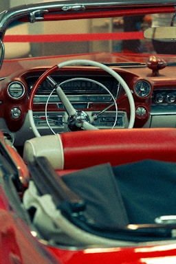 A vintage car's interior featuring a classic design with a red and white color scheme. The dashboard includes retro-style dials and a large steering wheel. The seats are upholstered in red leather with white accents. The overall setting includes soft lighting that enhances the nostalgic feel of the car.