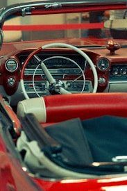 A vintage car's interior featuring a classic design with a red and white color scheme. The dashboard includes retro-style dials and a large steering wheel. The seats are upholstered in red leather with white accents. The overall setting includes soft lighting that enhances the nostalgic feel of the car.