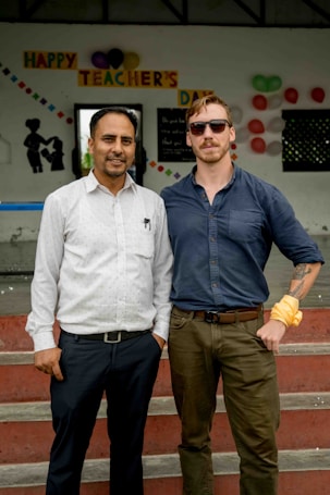 Two individuals are standing side by side on a set of steps in front of a wall with a 'Happy Teacher's Day' sign decorated with colorful paper letters and balloons. The person on the left is wearing a white shirt with a pen in the pocket, while the person on the right is wearing a blue shirt, sunglasses, and has a tattoo on the arm.