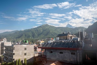 A cityscape with several mid-rise buildings in the foreground, featuring rooftops with tanks and solar panels. In the background, a lush green mountain range under a sky with scattered clouds. A snow-capped peak is also visible.