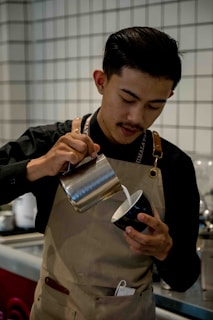 A barista wearing a black shirt and brown apron is pouring milk from a metal pitcher into a black cup, possibly preparing a coffee drink. The background consists of a tiled wall and some coffee equipment.