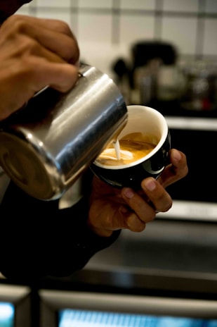 A person is pouring steamed milk from a pitcher into a cup of espresso, creating a latte art pattern. The hands are holding the pitcher and the cup delicately, showcasing the skill involved in latte preparation. The background includes blurred elements of a kitchen or cafe setting.