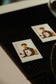 Two instant photos are placed on a textured black surface. Each photo contains an image of a person sitting in a casual indoor setting with a blurred background featuring light-colored walls and some shelves.