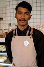 A person wearing a black shirt and beige apron stands in front of a display case filled with baked goods. The background features a tiled wall and stacked cups.