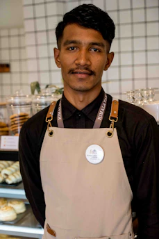 A person wearing a black shirt and beige apron stands in front of a display case filled with baked goods. The background features a tiled wall and stacked cups.