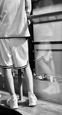 A person is wearing a casual, sporty outfit consisting of white shorts, a matching top, and sneakers. They are standing on a wooden floor next to a railing. The setting appears to be urban, with reflections in glass panels suggesting buildings or a cityscape.