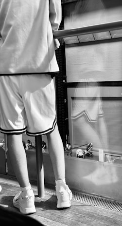 A person is wearing a casual, sporty outfit consisting of white shorts, a matching top, and sneakers. They are standing on a wooden floor next to a railing. The setting appears to be urban, with reflections in glass panels suggesting buildings or a cityscape.