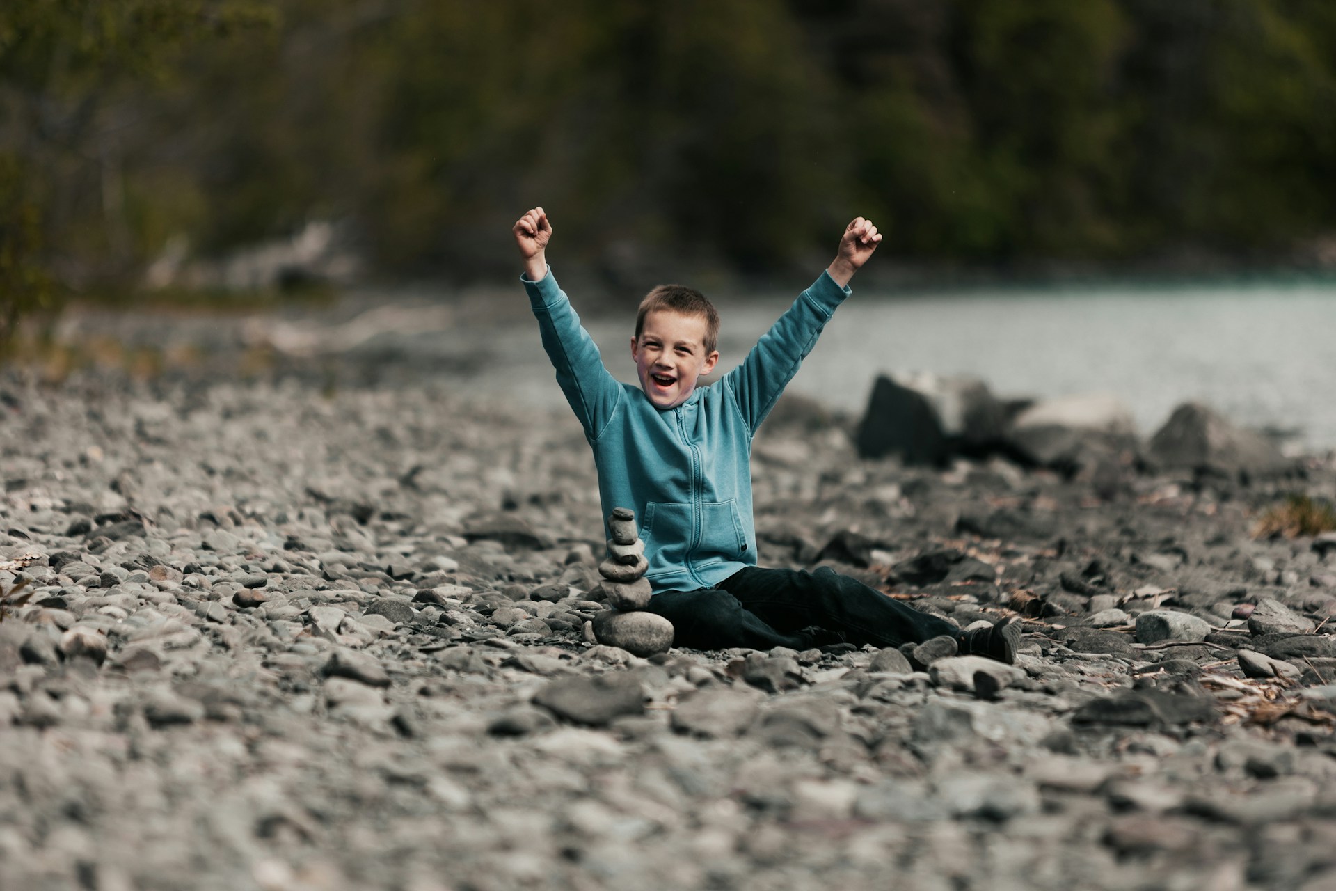 A young child with a joyful expression sits on a rocky shore, wearing a light blue jacket and black pants. They have their arms raised in triumph, and a small stack of balanced stones is placed in front of them. The background includes a blurred view of water and dense greenery, suggesting a natural and serene setting.