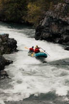 A group of adventurers navigate a rapid river in a bright blue inflatable raft. They are equipped with helmets and life vests, paddling with yellow oars as they maneuver through the white water. The surrounding landscape features rugged rocks and lush greenery.