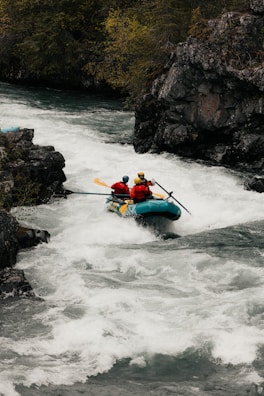 A group of adventurers navigate a rapid river in a bright blue inflatable raft. They are equipped with helmets and life vests, paddling with yellow oars as they maneuver through the white water. The surrounding landscape features rugged rocks and lush greenery.