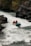A group of adventurers navigate a rapid river in a bright blue inflatable raft. They are equipped with helmets and life vests, paddling with yellow oars as they maneuver through the white water. The surrounding landscape features rugged rocks and lush greenery.
