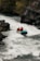 A group of adventurers navigate a rapid river in a bright blue inflatable raft. They are equipped with helmets and life vests, paddling with yellow oars as they maneuver through the white water. The surrounding landscape features rugged rocks and lush greenery.