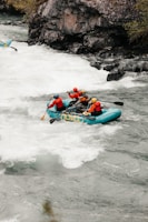 A group of four people is navigating through the rapids in a teal inflatable raft. They are all wearing safety gear, including helmets and life jackets. The river is turbulent with white water, and there are rocky cliffs and vegetation along the riverbank.