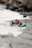 A group of four people is navigating through the rapids in a teal inflatable raft. They are all wearing safety gear, including helmets and life jackets. The river is turbulent with white water, and there are rocky cliffs and vegetation along the riverbank.