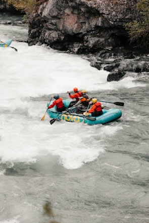 A group of four people is navigating through the rapids in a teal inflatable raft. They are all wearing safety gear, including helmets and life jackets. The river is turbulent with white water, and there are rocky cliffs and vegetation along the riverbank.
