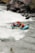 A group of four people is navigating through the rapids in a teal inflatable raft. They are all wearing safety gear, including helmets and life jackets. The river is turbulent with white water, and there are rocky cliffs and vegetation along the riverbank.